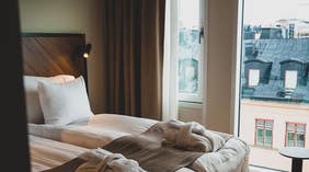 En bäddad dubbelsäng med morgonrockar i fotändan och stora fönster med utsikt över takåsarna  i ett Deluxerum på Clarion Hotel Amaranten i Stockholm.